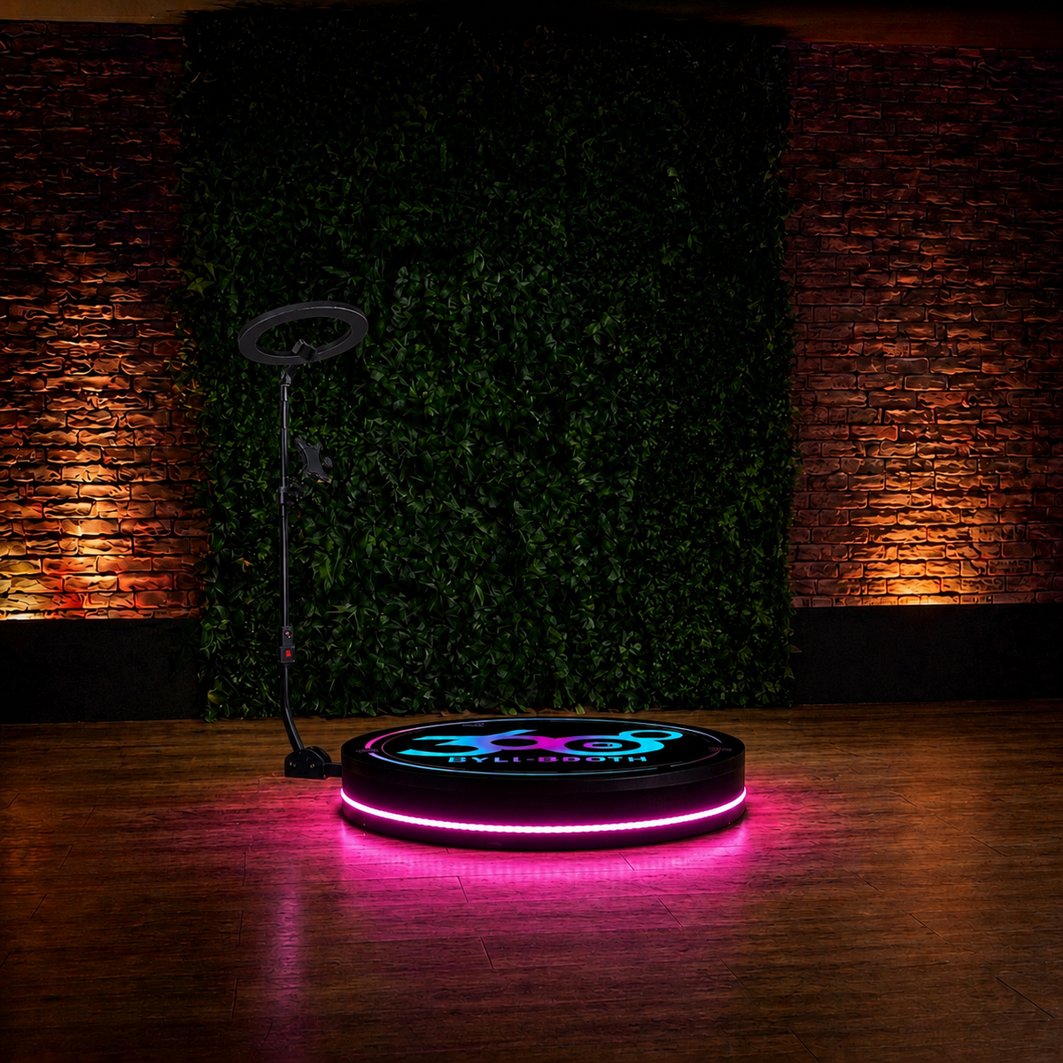 A round black LED 360 video booth with a glowing pink outer ring and a blue/pink logo in the center, set on a wooden floor in front of a green hedge wall with warm brick lighting.