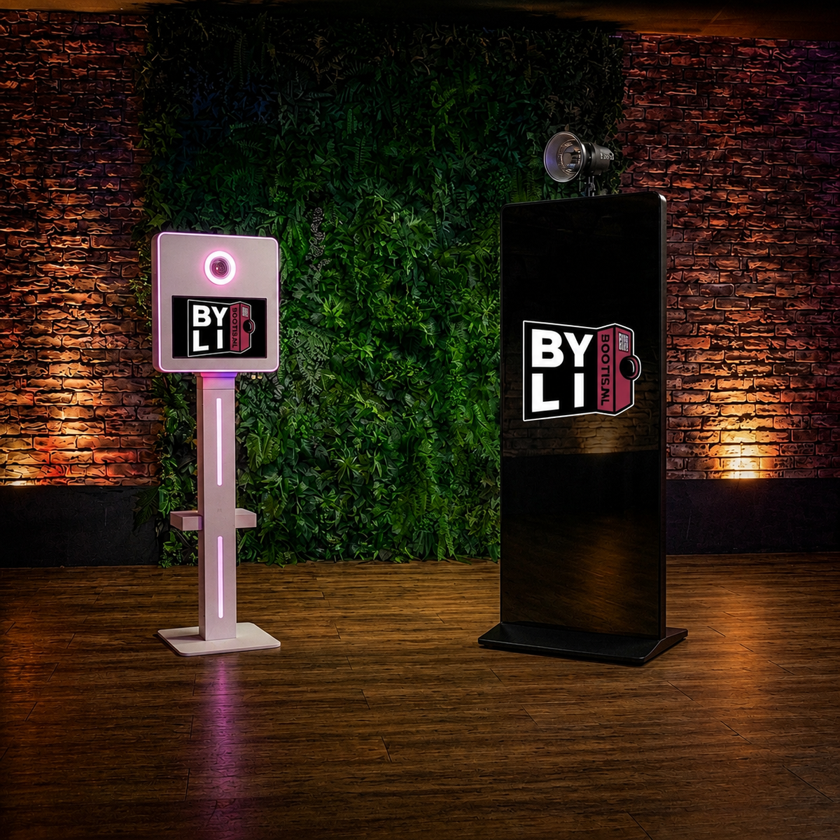 Two BY LI branded event photo booths on a wooden stage in front of a green wall and brick wall backdrop. One machine is a freestanding white face scanner with pink lighting, and the other is a tall black display stand with a screen showing the BY LI logo.
